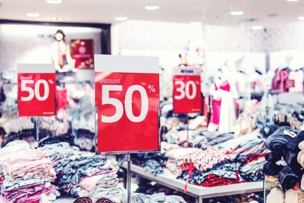 Clearance reselling Amazon - Red and white signs prominently display "50%" and "30%" discounts amidst racks and piles of clothing, representing the opportunity to find clearance items for reselling.