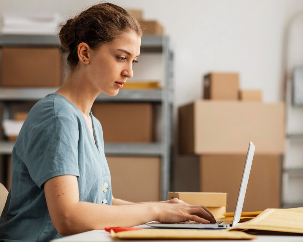 Amazon messages - A woman working on a laptop amidst shipping materials, checking or responding to Amazon messages related to her business.
