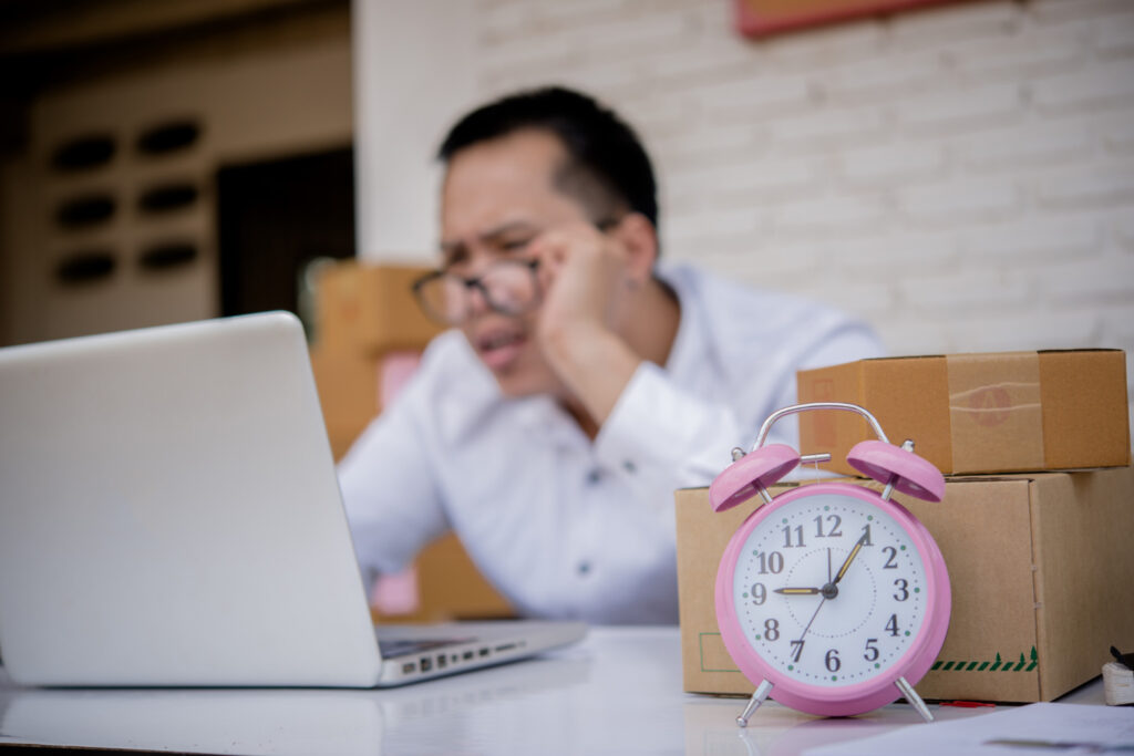 Amazon shipping delays - Stressed person at laptop with a pink alarm clock and packages.