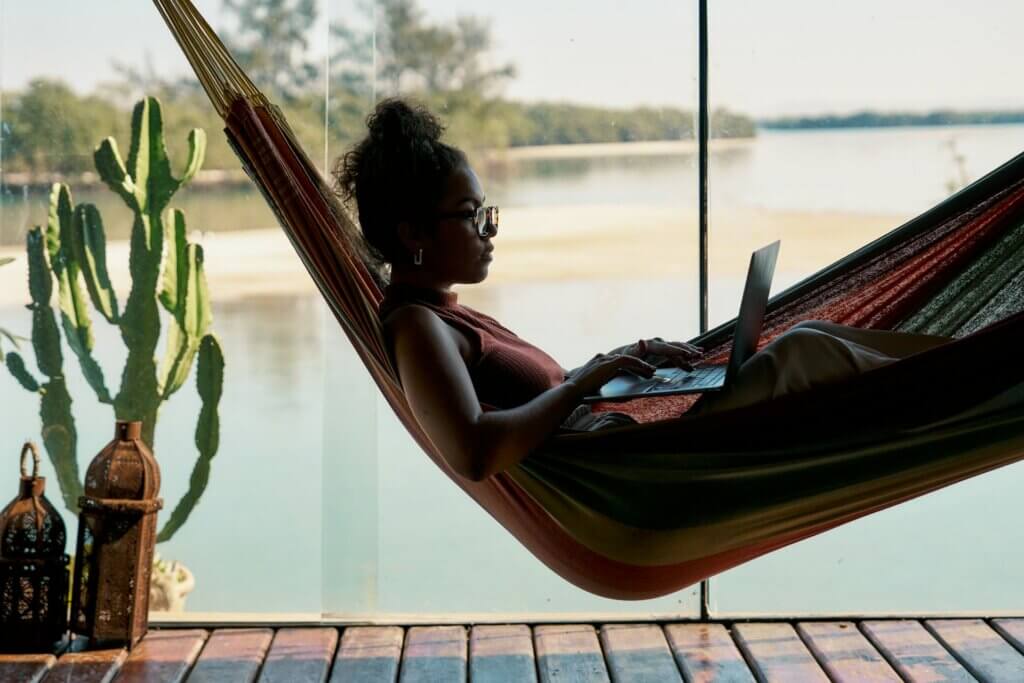 Become a digital nomad selling online - A woman working on a laptop while relaxing in a hammock by a scenic view