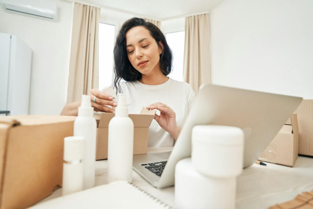 amazon fba new selection program guide - a woman sitting carefully packing white cosmetic bottles into a cardboard box for shipping